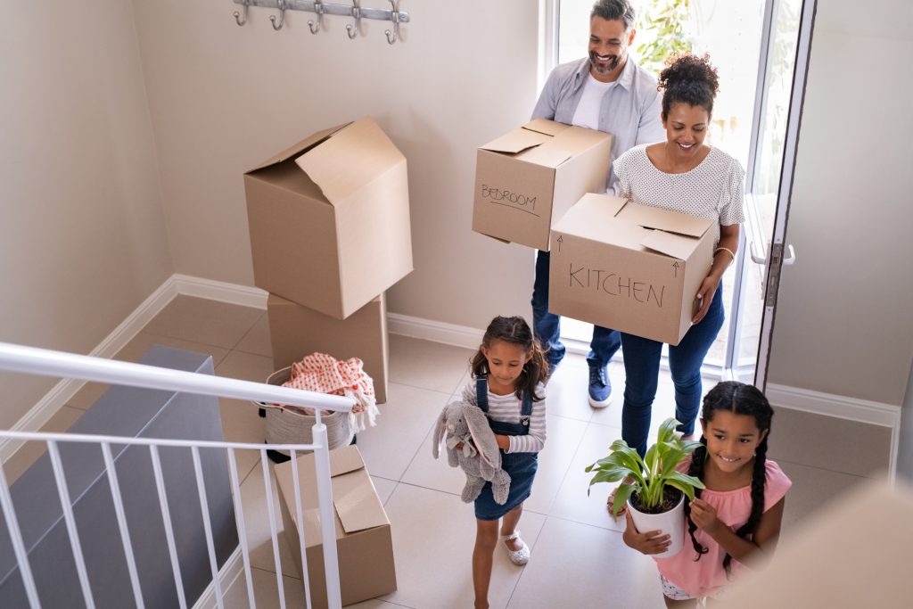 Ethnic family with two children carrying boxes and plant in new home on moving day. High angle view of happy smiling daughters helping mother and father with cardboard boxes in new house. Top view of excited kids having fun walking up stairs running to their rooms while parents holding boxes.