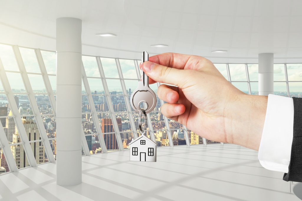 person's hand with suit on holding out a key to an apartment with windows in the background overlooking city