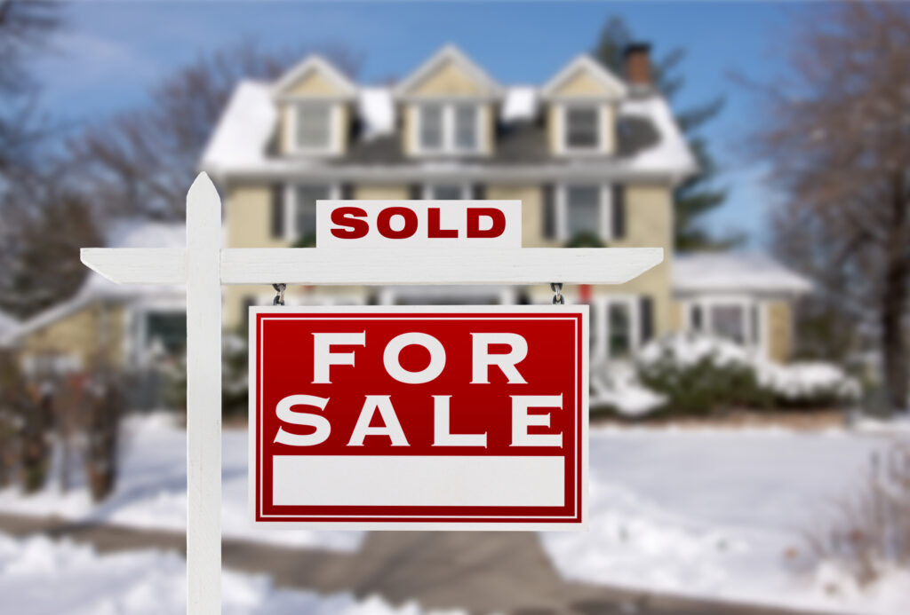 House in the background with snow on the ground around it. A forsale with a sold sign on it in the foreground.