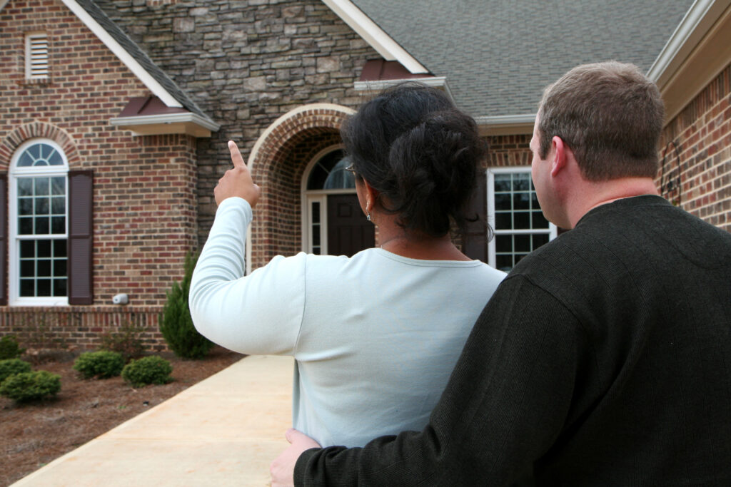 A view of a couple from behind as they loo at a house and point to something interesting about the brick house.