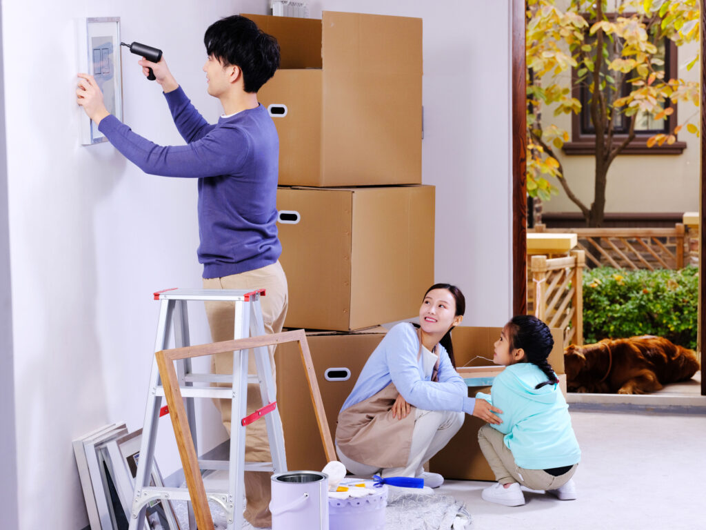 Man standing near a wall hanging artwork with woman and child on the floor nearby watching as they sit in front of stacked moving boxes.